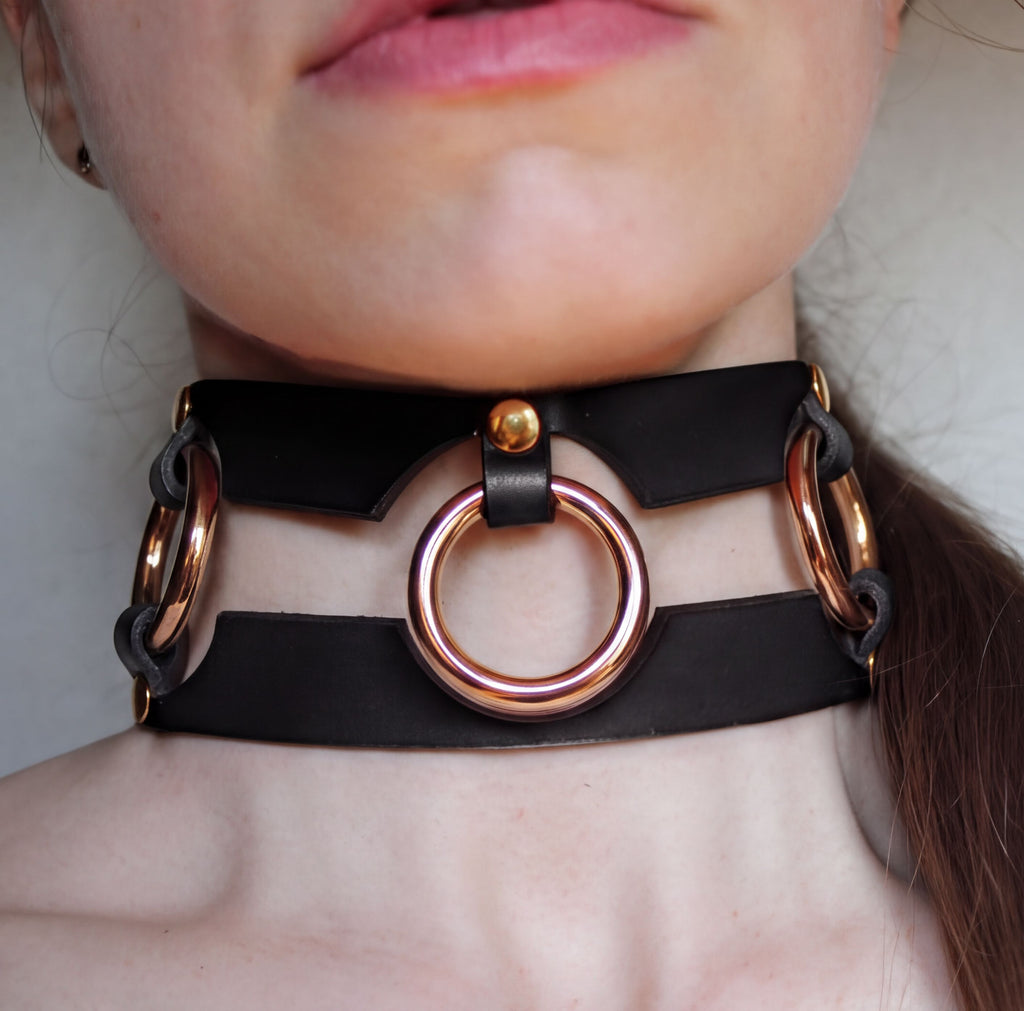 A close-up of a person's neck wearing a black leather choker necklace with three circular metal rings.