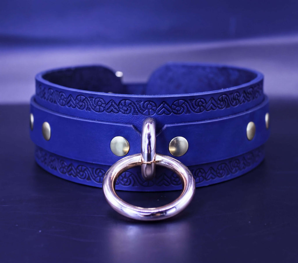 A blue leather choker necklace with a gold ring and metal studs, displayed on a dark surface.