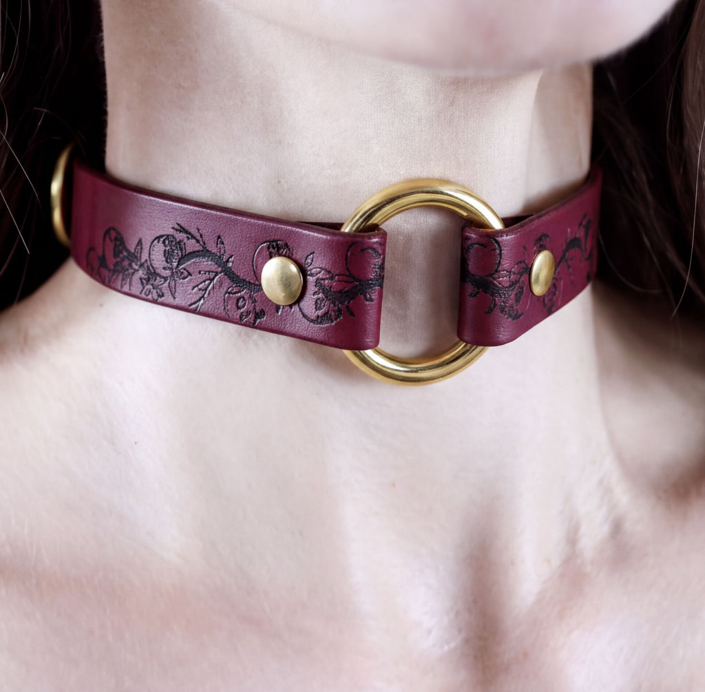 A close-up view of a burgundy leather choker necklace with a gold ring and two gold-colored buttons.