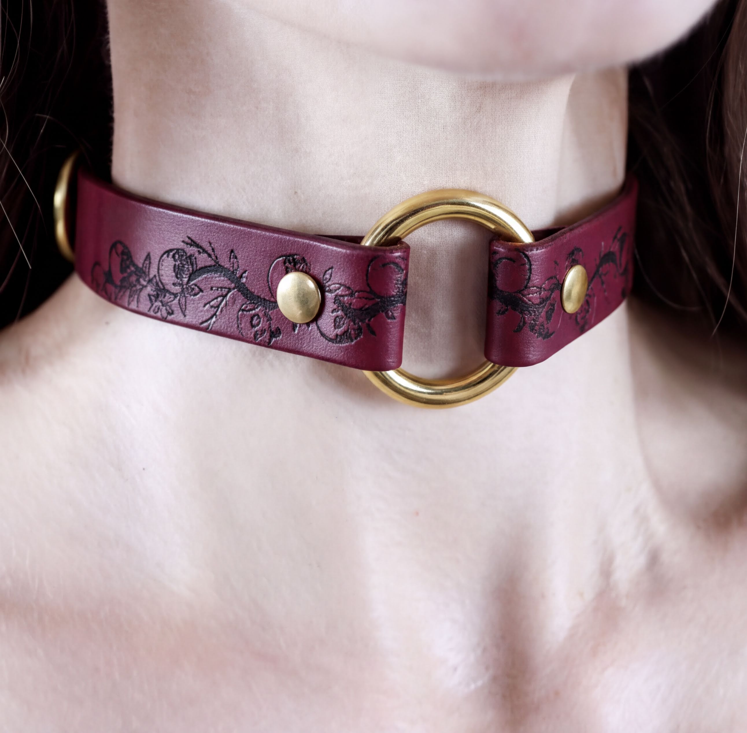 A close-up view of a burgundy leather choker necklace with a gold ring and two gold-colored buttons.