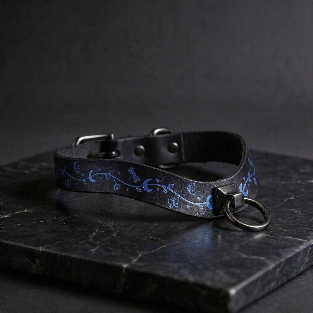 A black leather collar with a blue floral design and a metal ring on the front, resting on a dark surface.