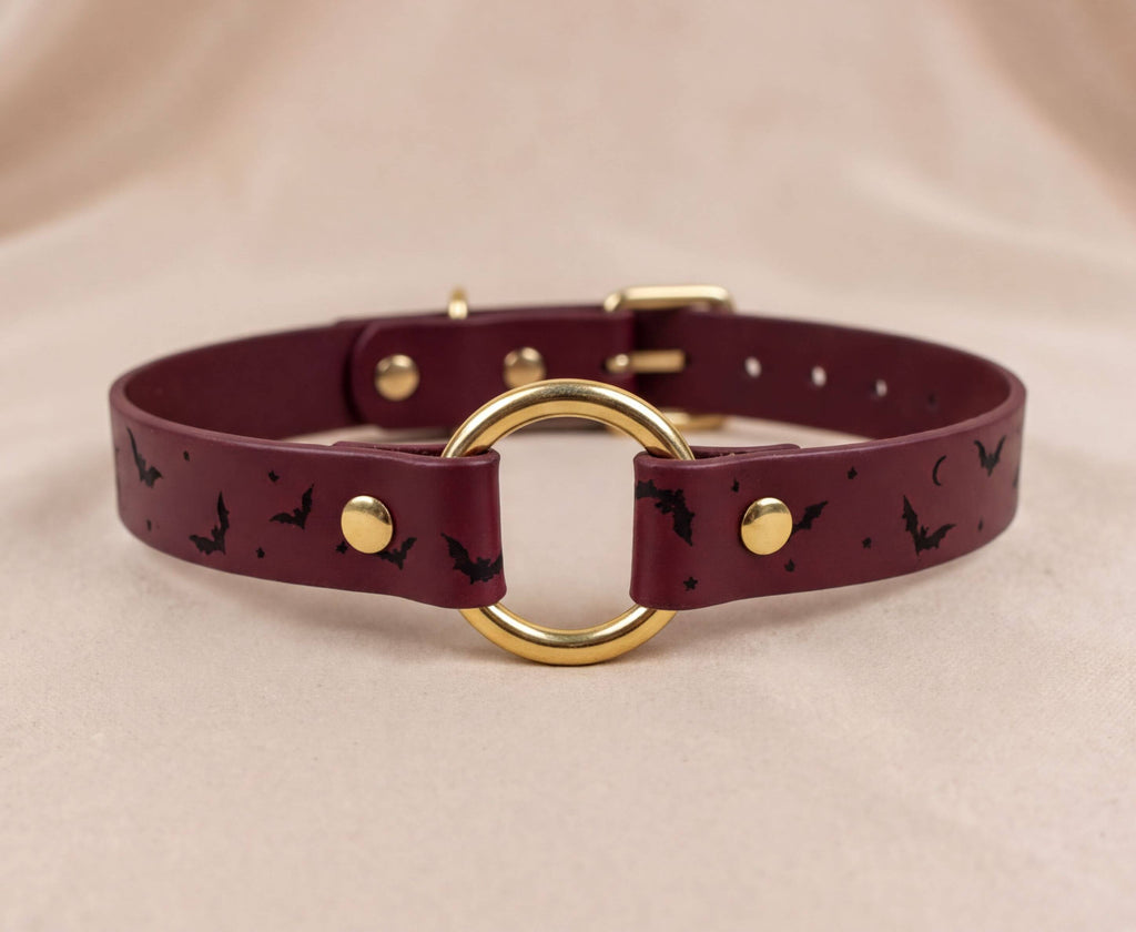 A burgundy leather collar with a gold ring and black bat designs.