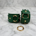a pair of green leather cuffs with gold hardware, including two large rings and a smaller ring, placed on a marble surface.