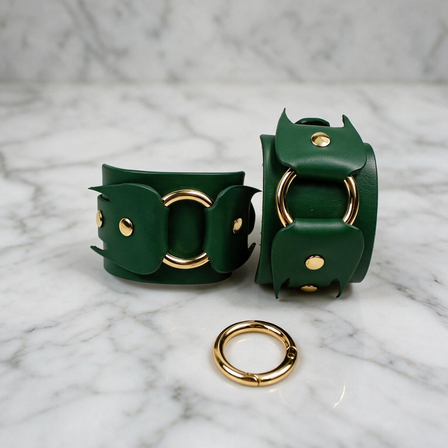 a pair of green leather cuffs with gold hardware, including two large rings and a smaller ring, placed on a marble surface.