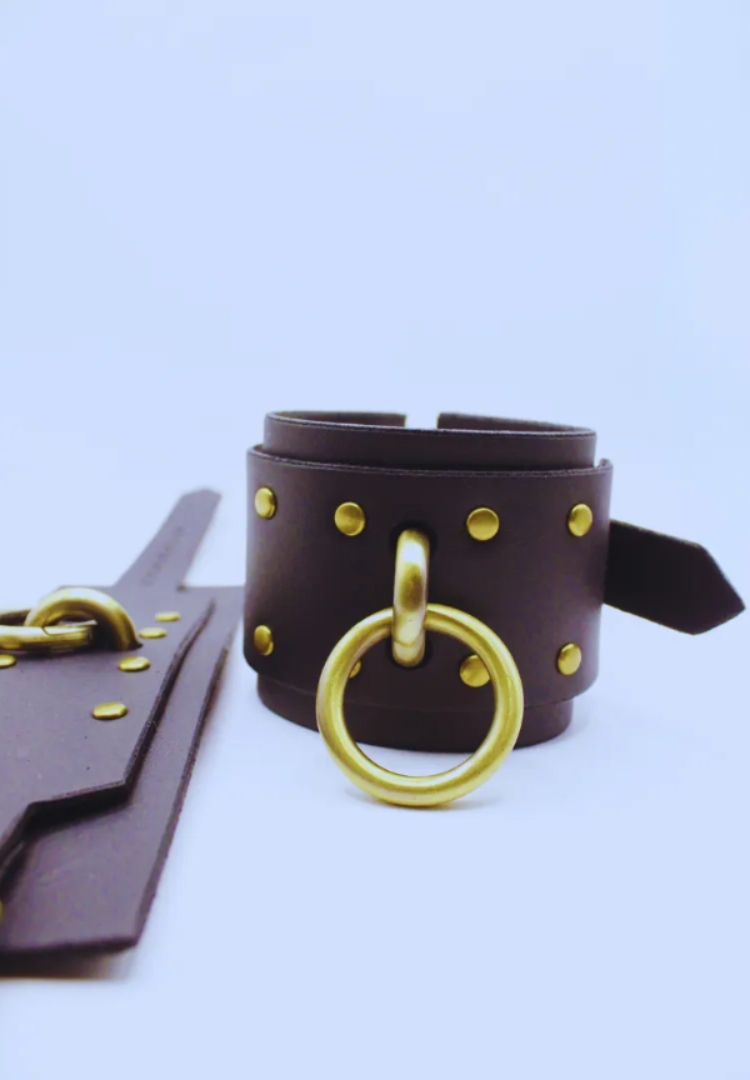 A dark brown leather strap with gold-colored metal rings and buckles, placed on a white background.