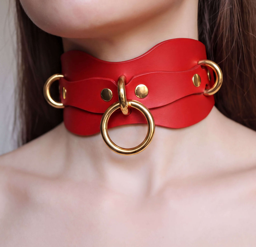 A close-up of a person's neck wearing a red leather choker necklace with gold-colored metal rings.