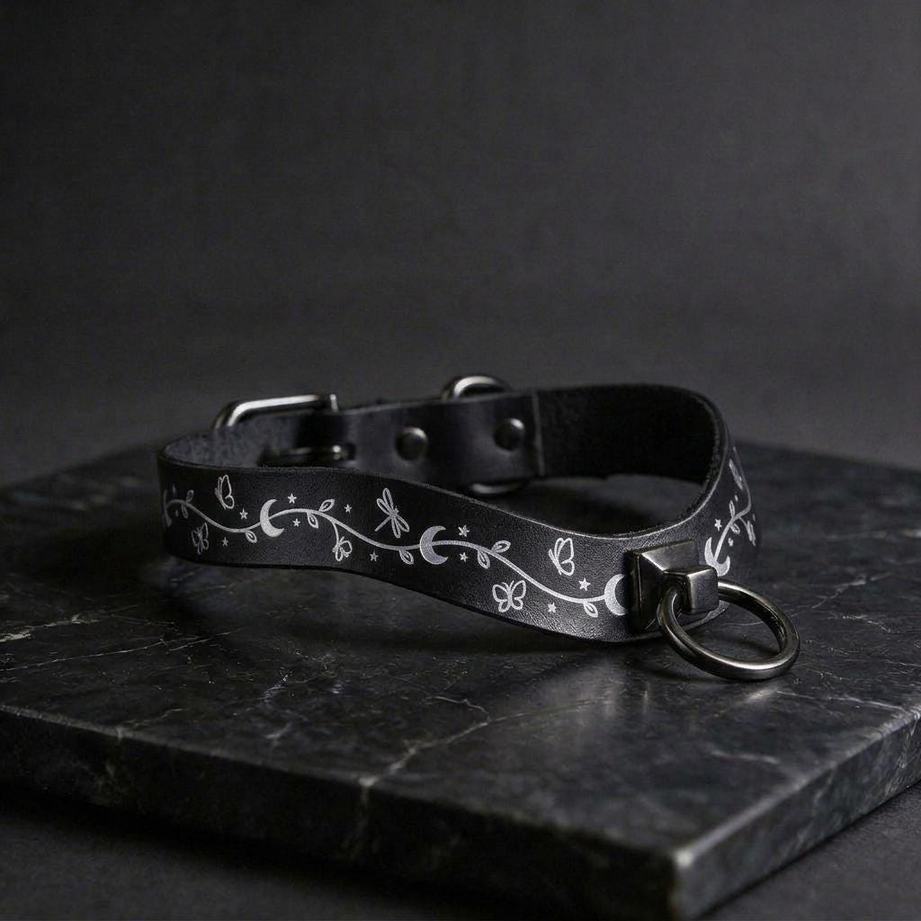 A black leather collar with a silver metal ring and a floral design on it, resting on a dark surface.