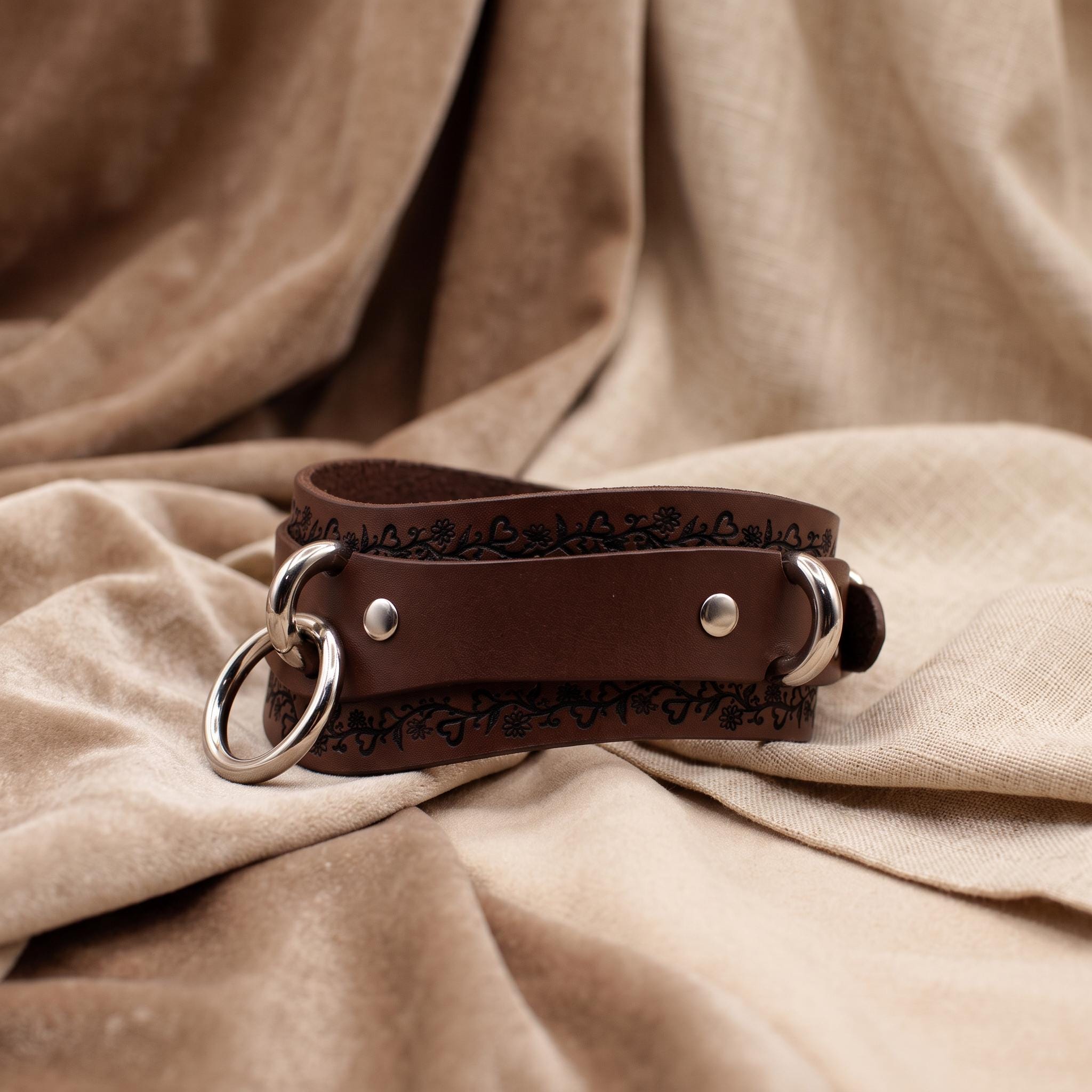 A brown leather collar with metal rings and a decorative pattern on the leather strap is displayed on a beige fabric background.