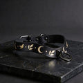 A black leather collar with a silver metal ring and a decorative pattern of leaves and crescent moons on it, resting on a dark surface.