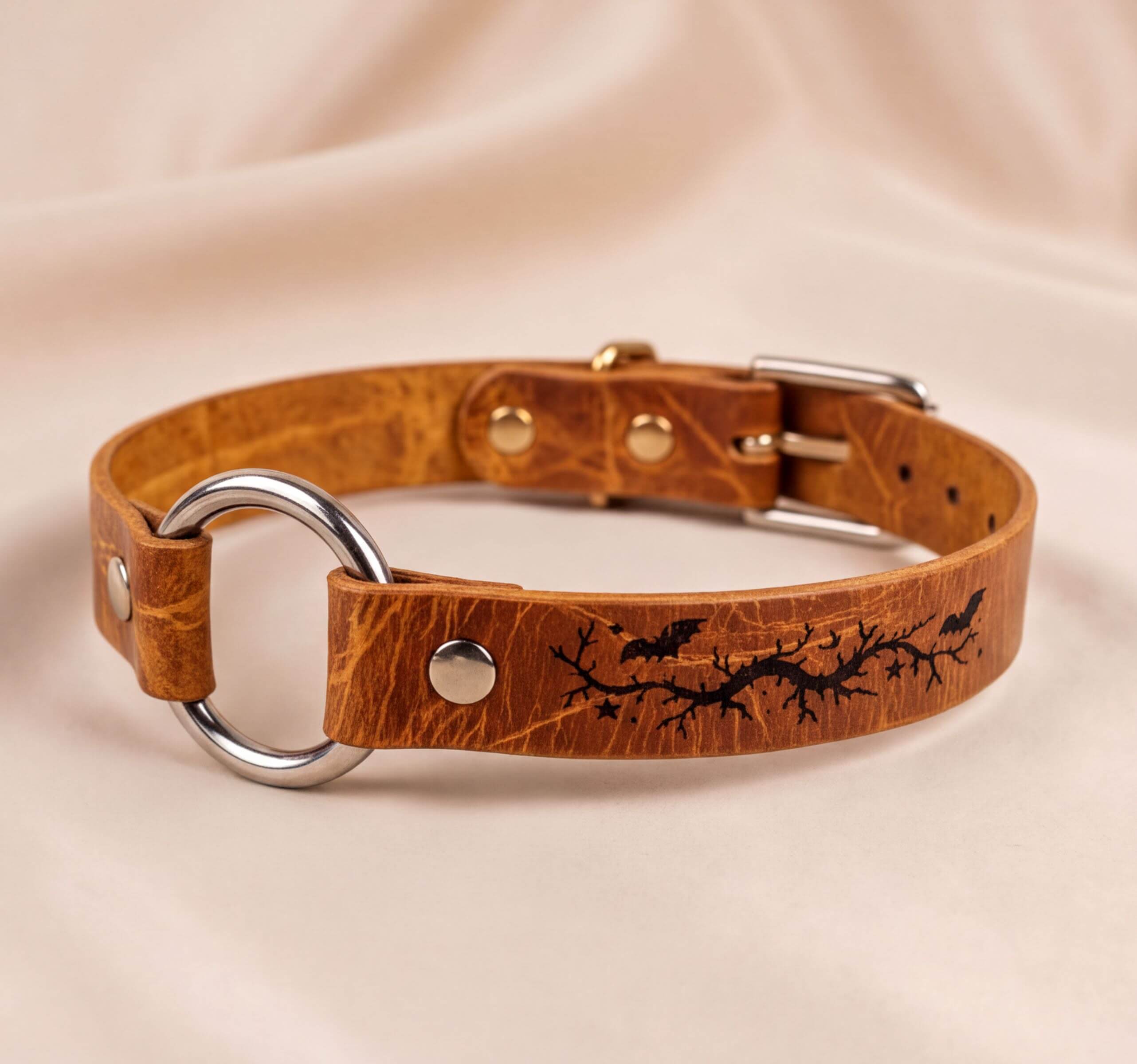 A brown leather collar with a metal ring and a design featuring bats and branches.