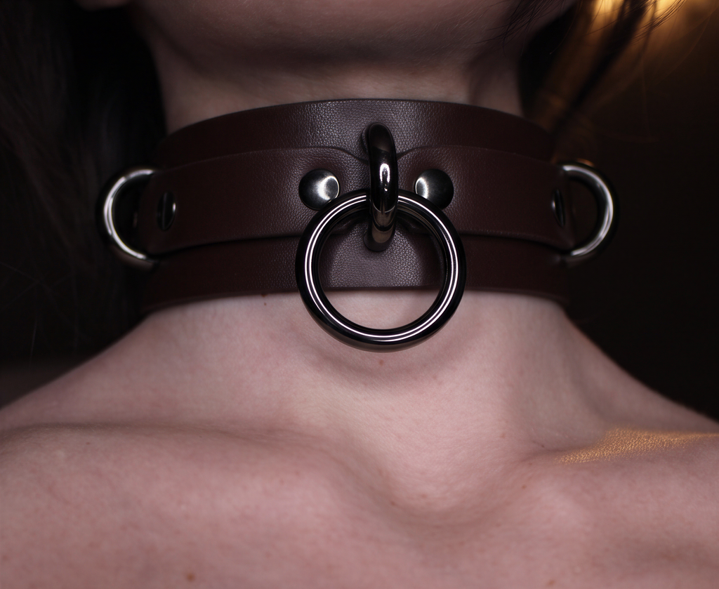 A close-up of a person's neck wearing a leather choker with metal rings and a metal clasp.