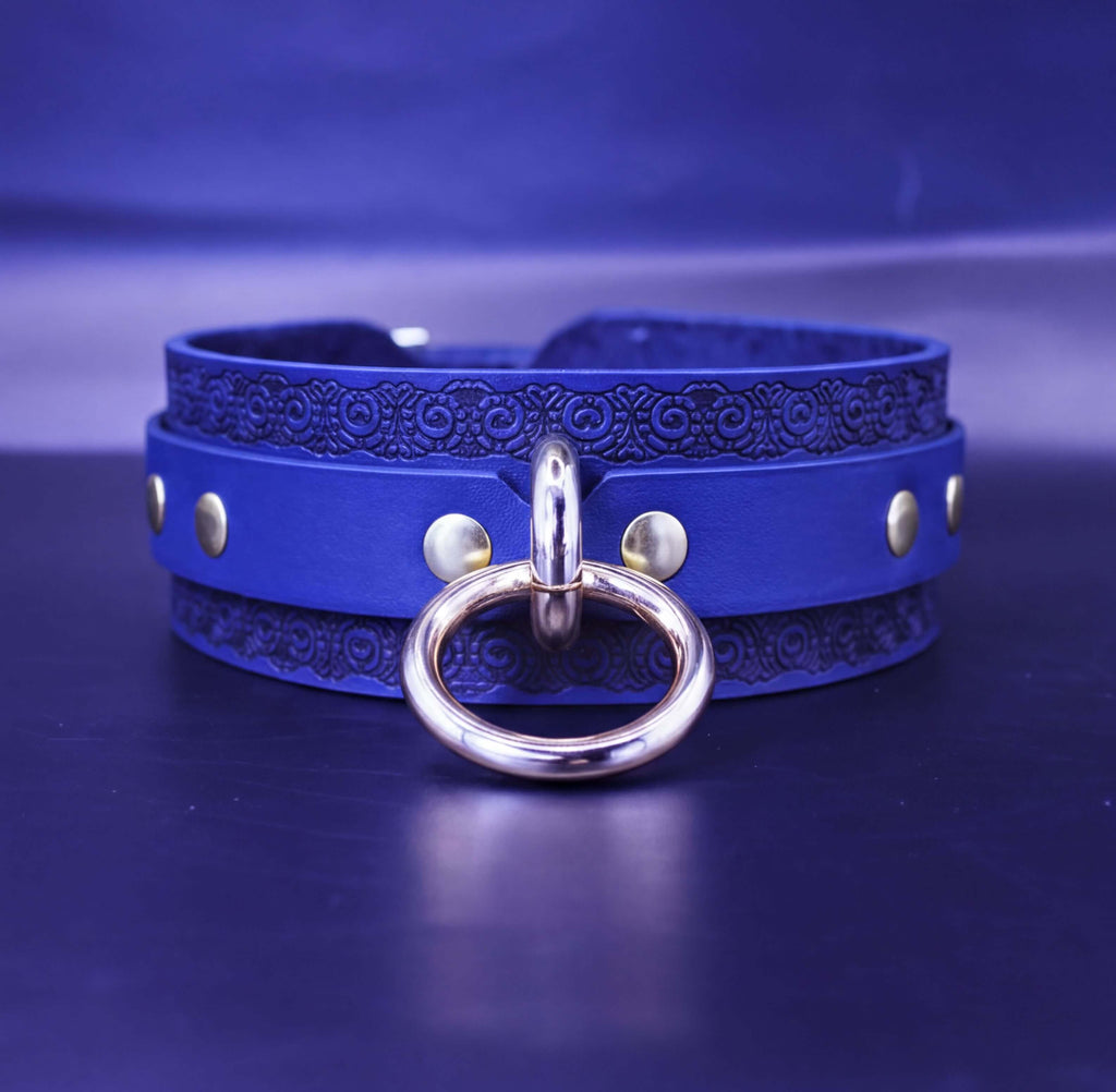 A blue leather choker necklace with a gold-colored metal ring and three silver-colored studs on the front.