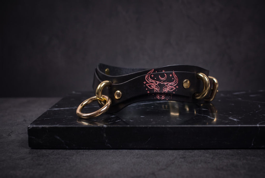 A black leather choker necklace with gold-colored metal rings and a red design on the clasp is displayed on a black marble surface.