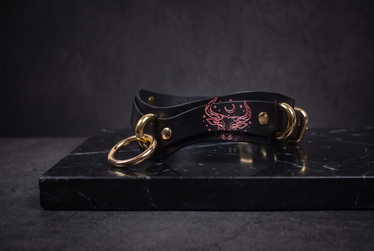 A black leather choker necklace with gold-colored metal rings and a red design on the clasp is displayed on a black marble surface.