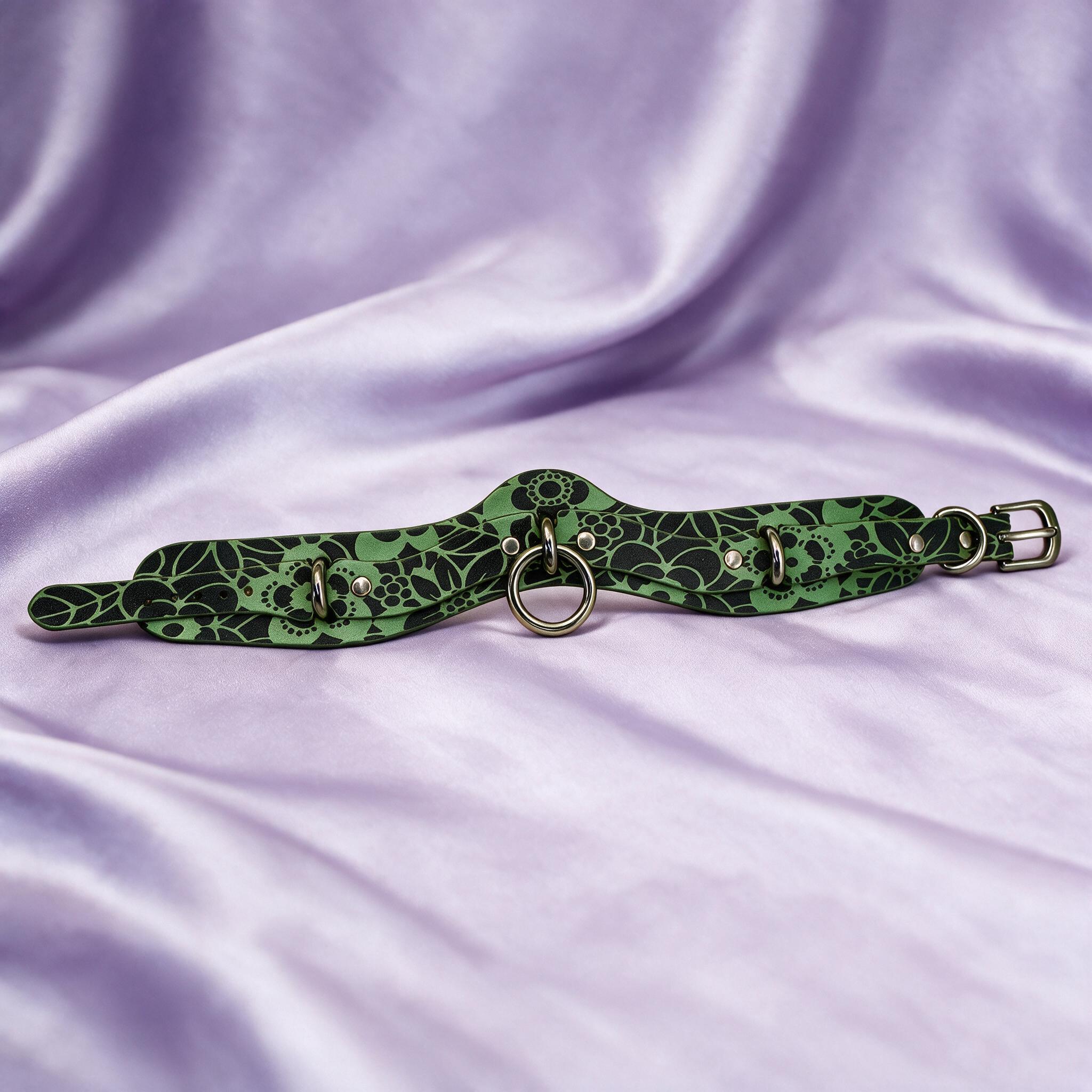 A green and black floral choker necklace with metal rings and a buckle is displayed on a purple fabric background.