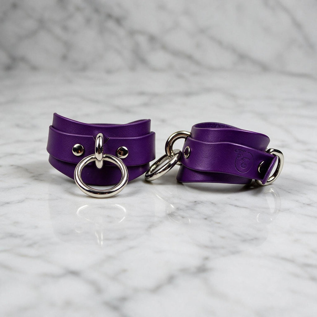 A pair of purple leather cuffs with metal rings and buckles, placed on a marble surface.