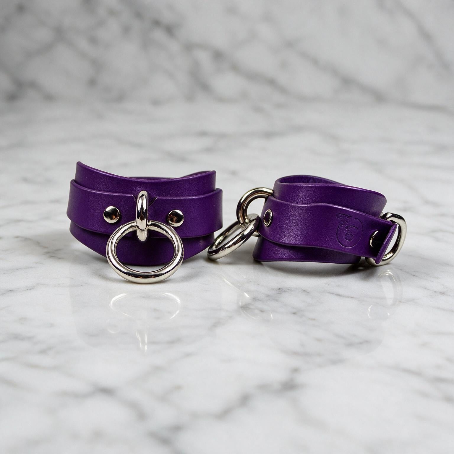 A pair of purple leather cuffs with metal rings and buckles, placed on a marble surface.