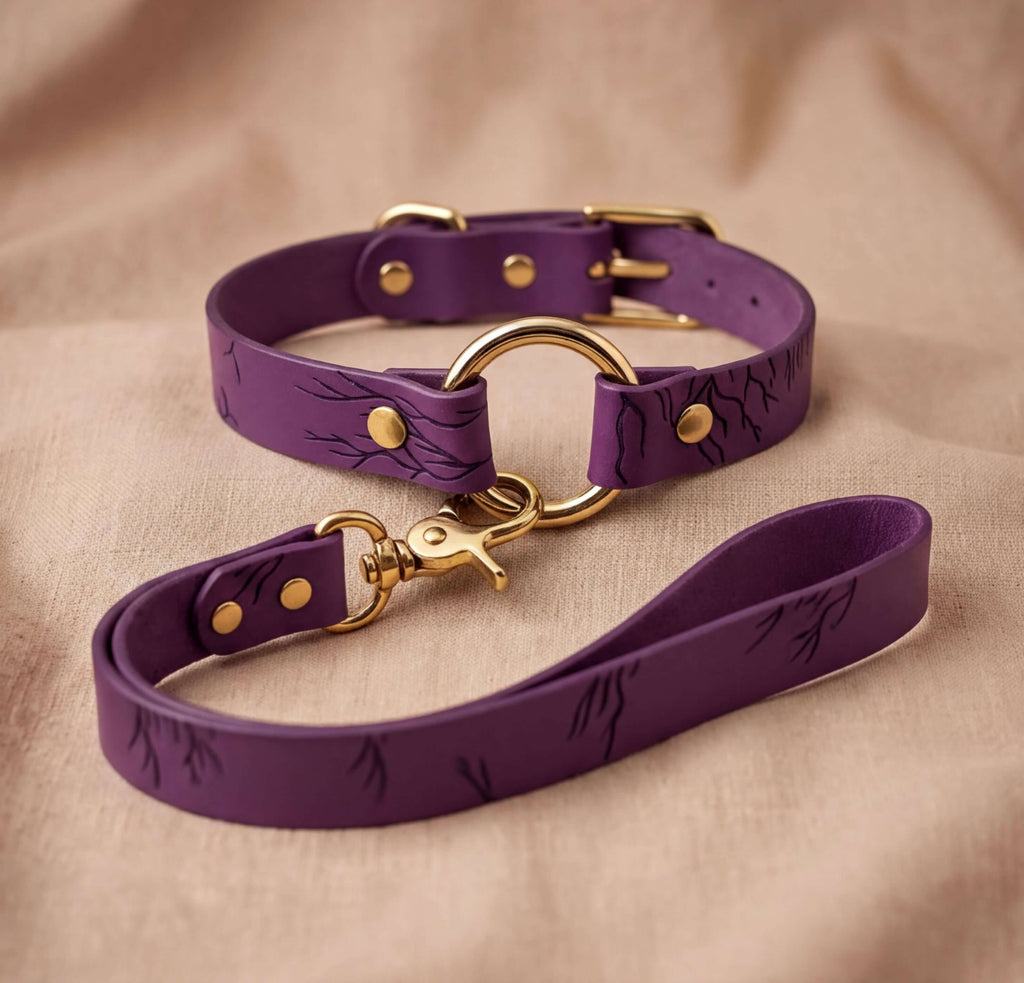 A purple leather dog collar with a gold-colored buckle and a matching leash is displayed on a beige fabric background.