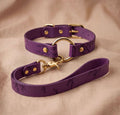 A purple leather dog collar with a gold-colored buckle and a matching leash is displayed on a beige fabric background.
