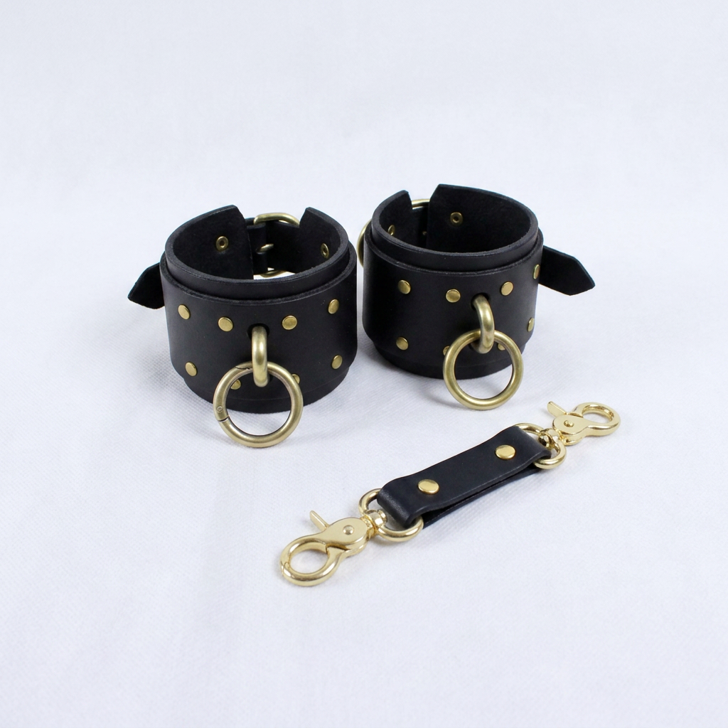 a pair of black leather cuffs with gold rings and a black leather strap with gold rings, all placed on a white background.