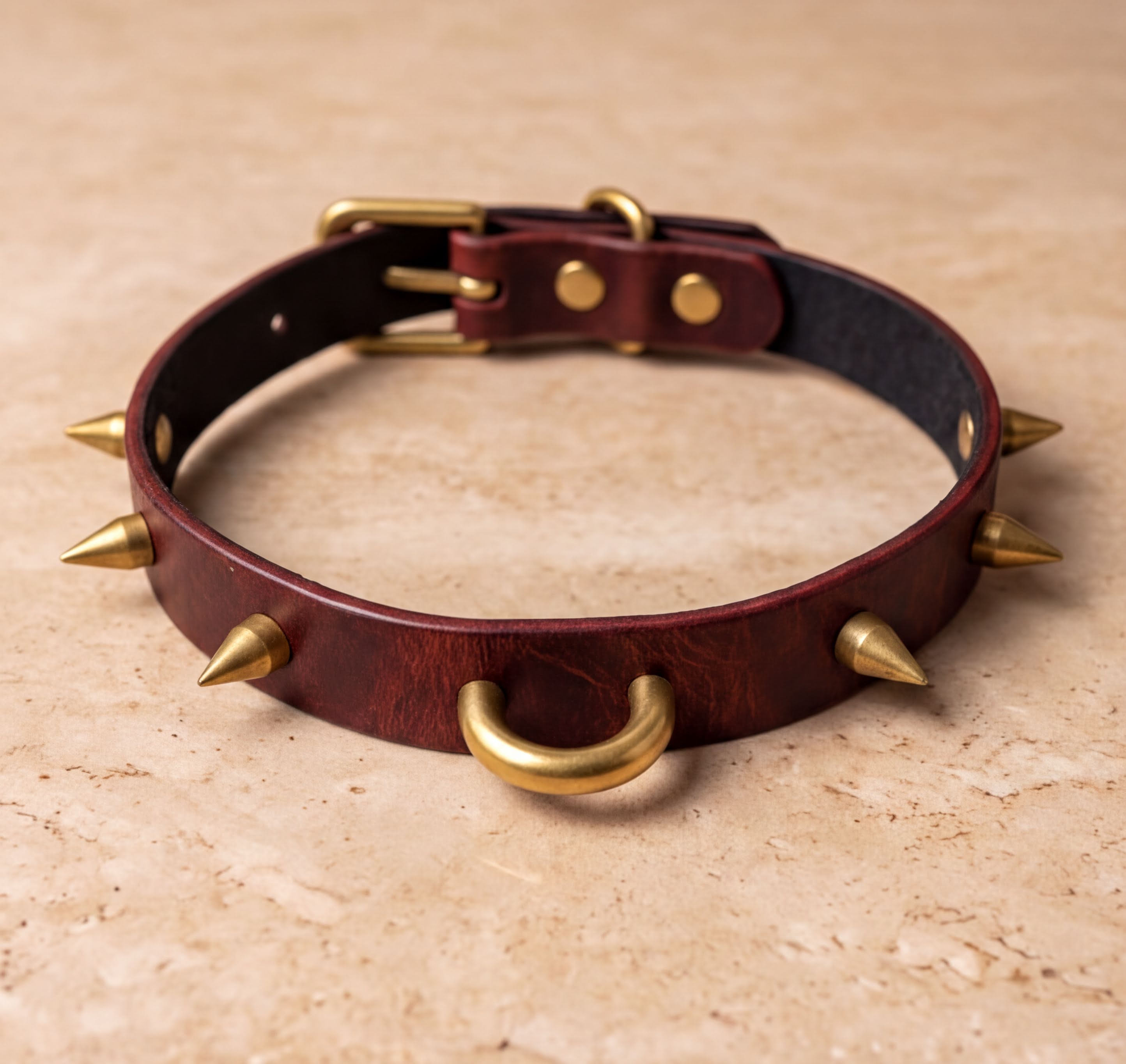 A burgundy leather collar with gold spikes protruding from it, resting on a beige surface.