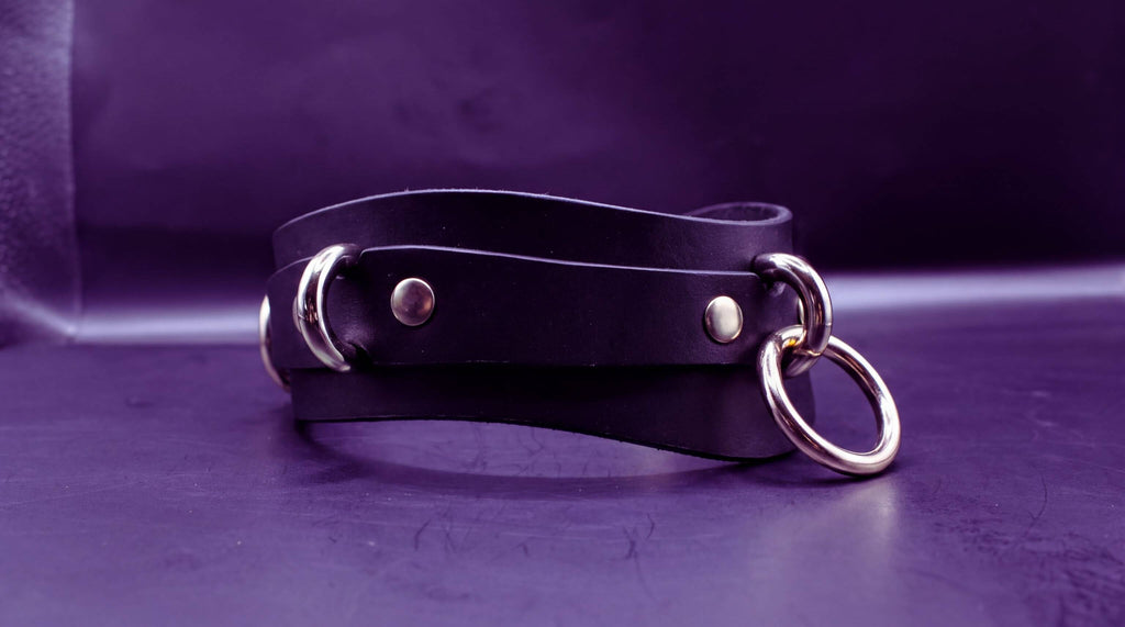 A black leather choker necklace with metal rings and buckles is displayed against a purple background.