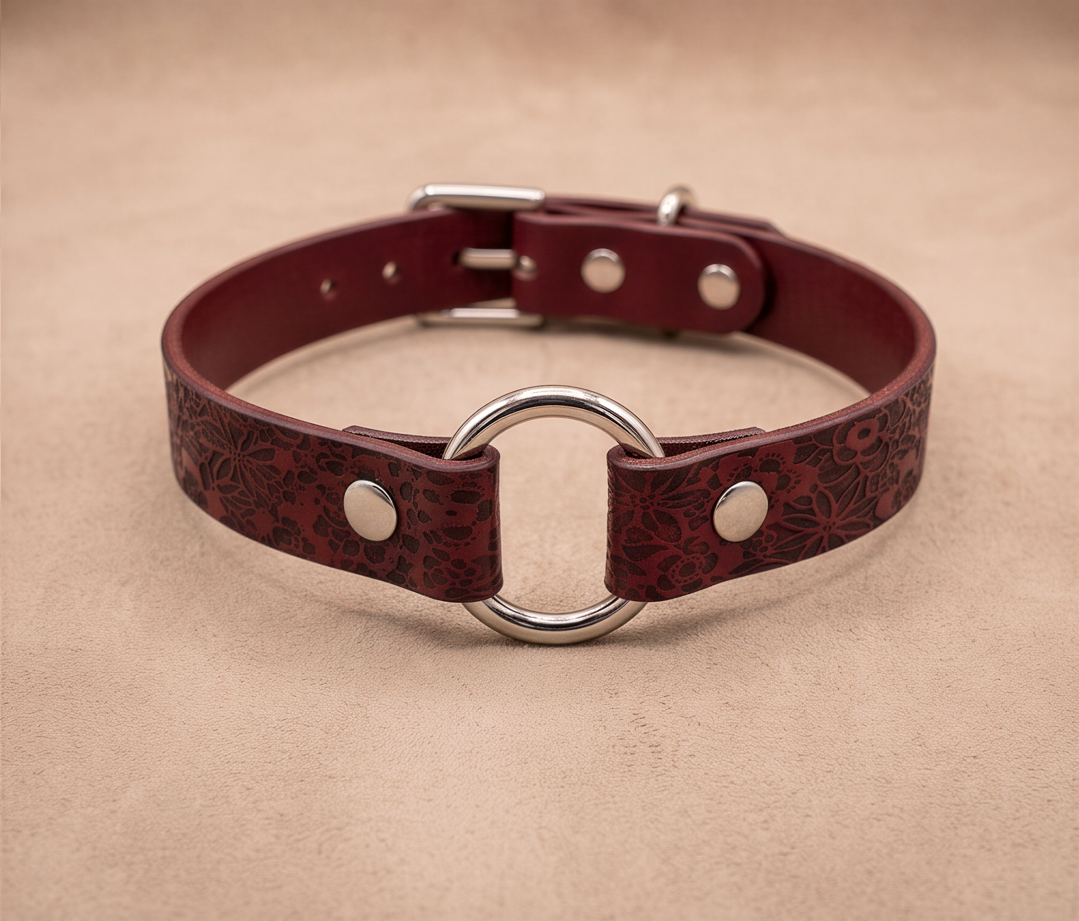A burgundy leather collar with a metal ring and buckle, displayed on a beige background.