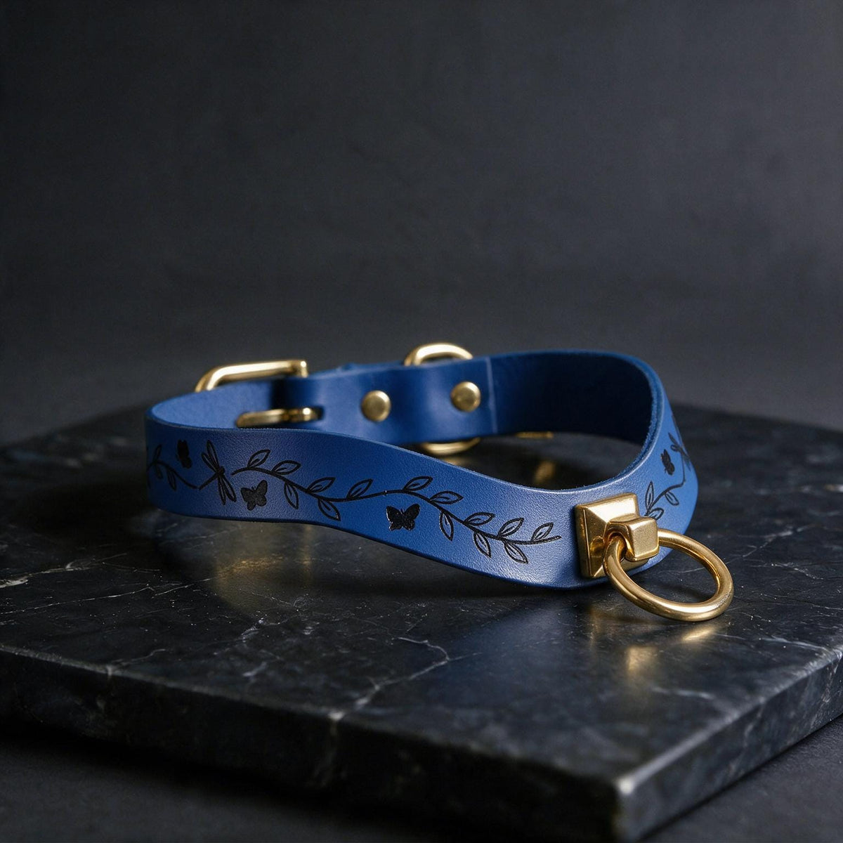 A blue leather collar with a leaf and butterfly design, featuring a gold-colored metal ring and buckle, is displayed on a black marble surface.