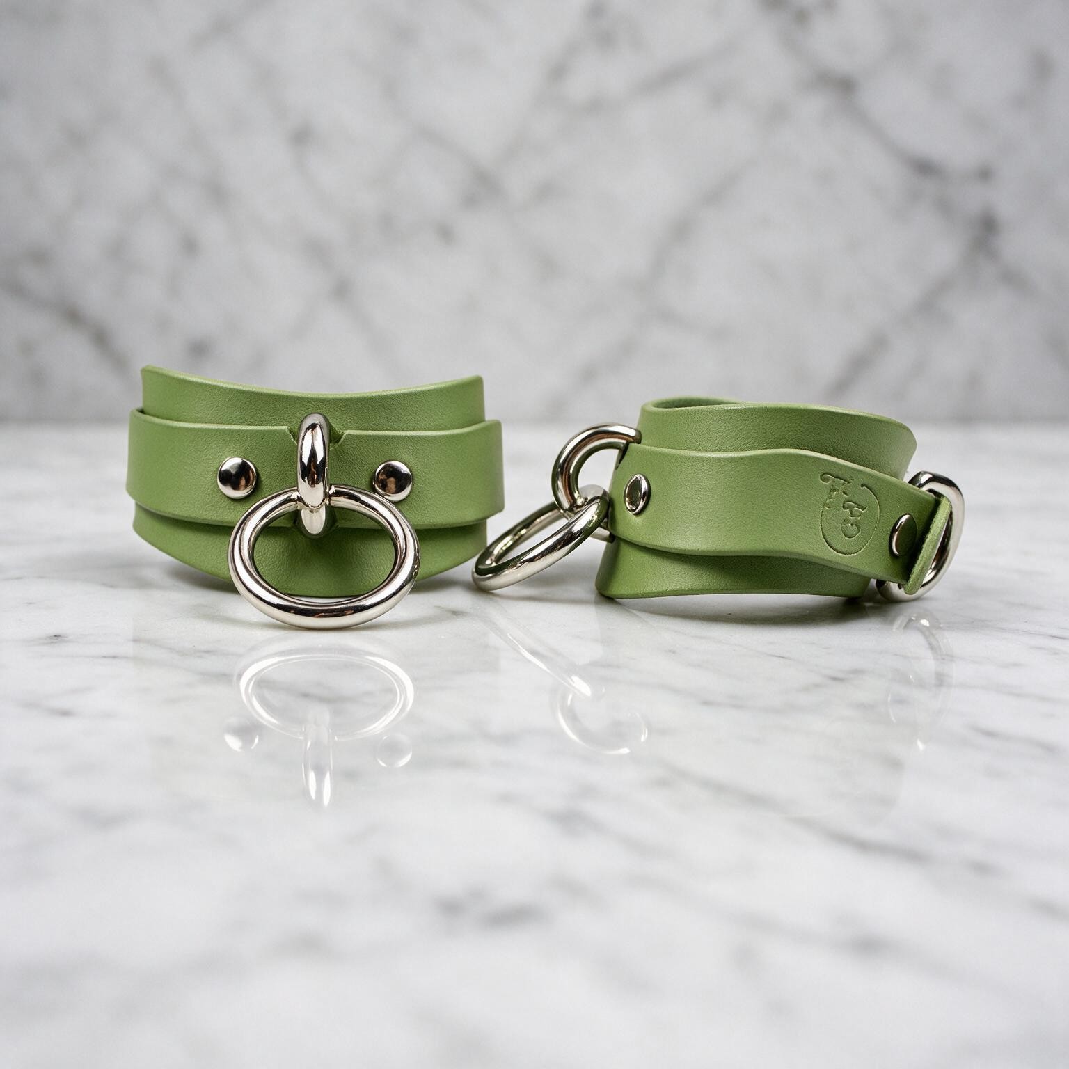 Two green leather cinch belts with metal rings and buckles are displayed on a marble surface.