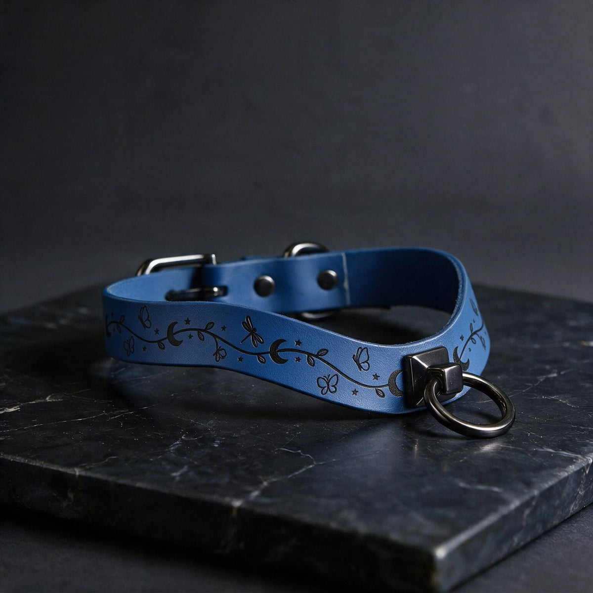 A blue leather collar with a metal ring and decorative elements rests on a dark surface.