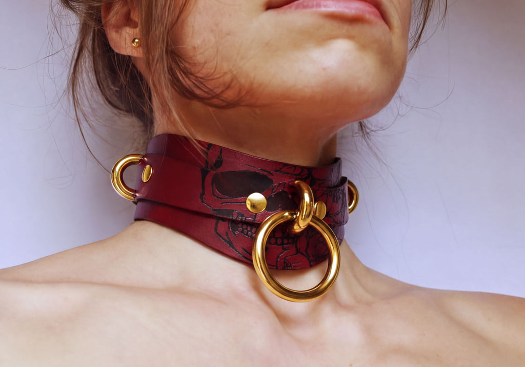 A close-up of a woman's neck and chest area wearing a burgundy leather choker necklace with gold-colored rings and a skull design.