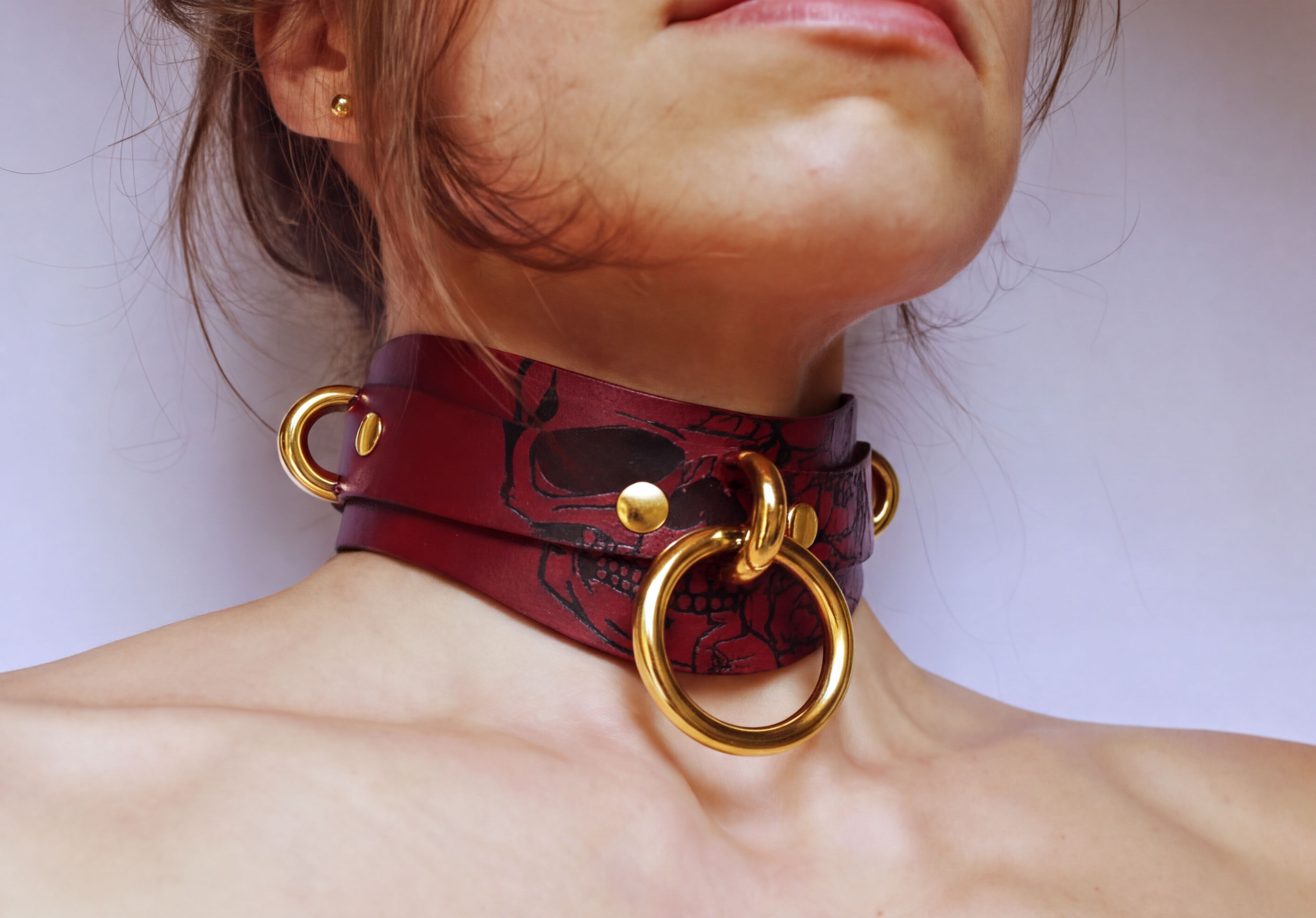 A close-up of a woman's neck and chest area wearing a burgundy leather choker necklace with gold-colored rings and a skull design.