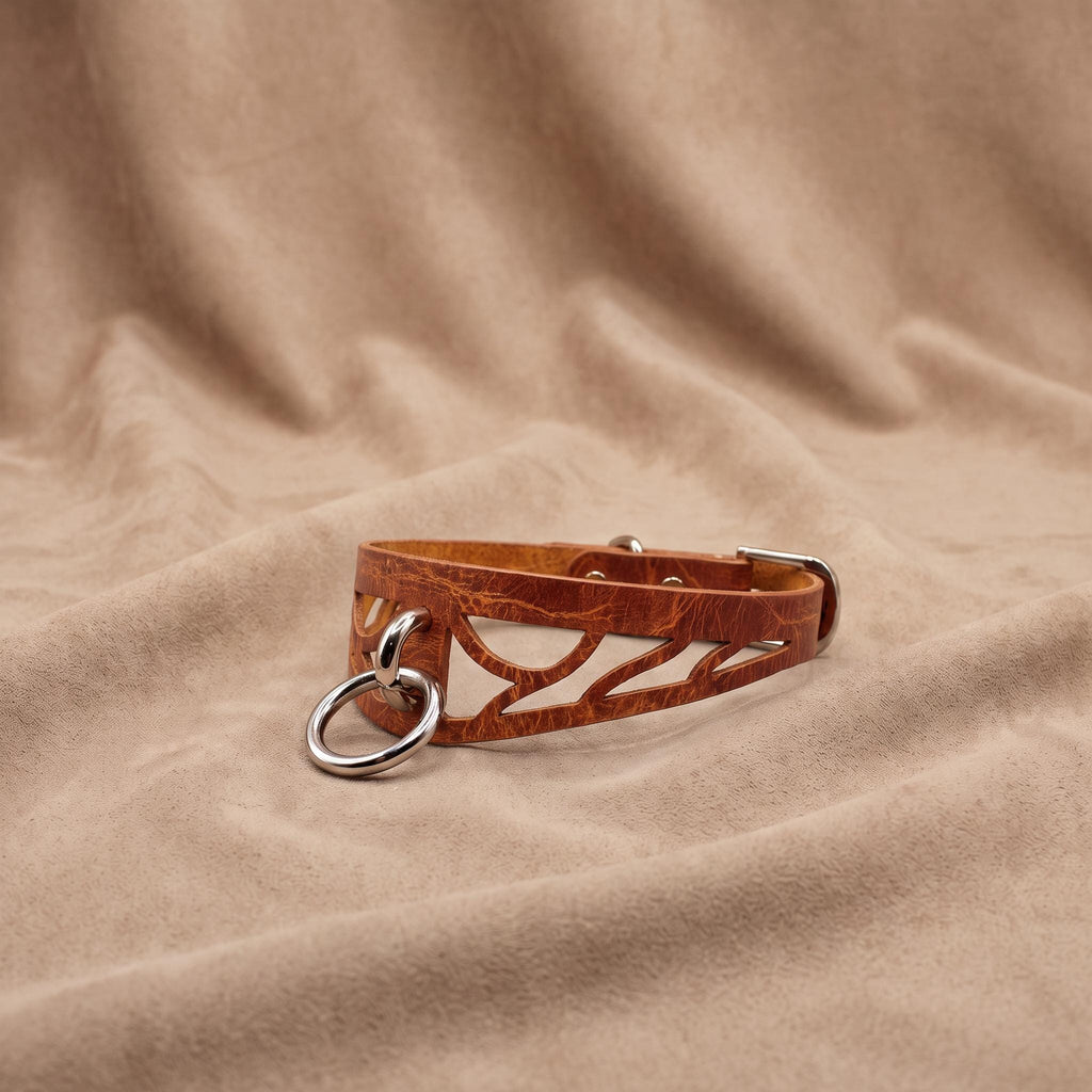 A brown leather choker necklace with a metal ring and buckle is displayed on a beige fabric background.
