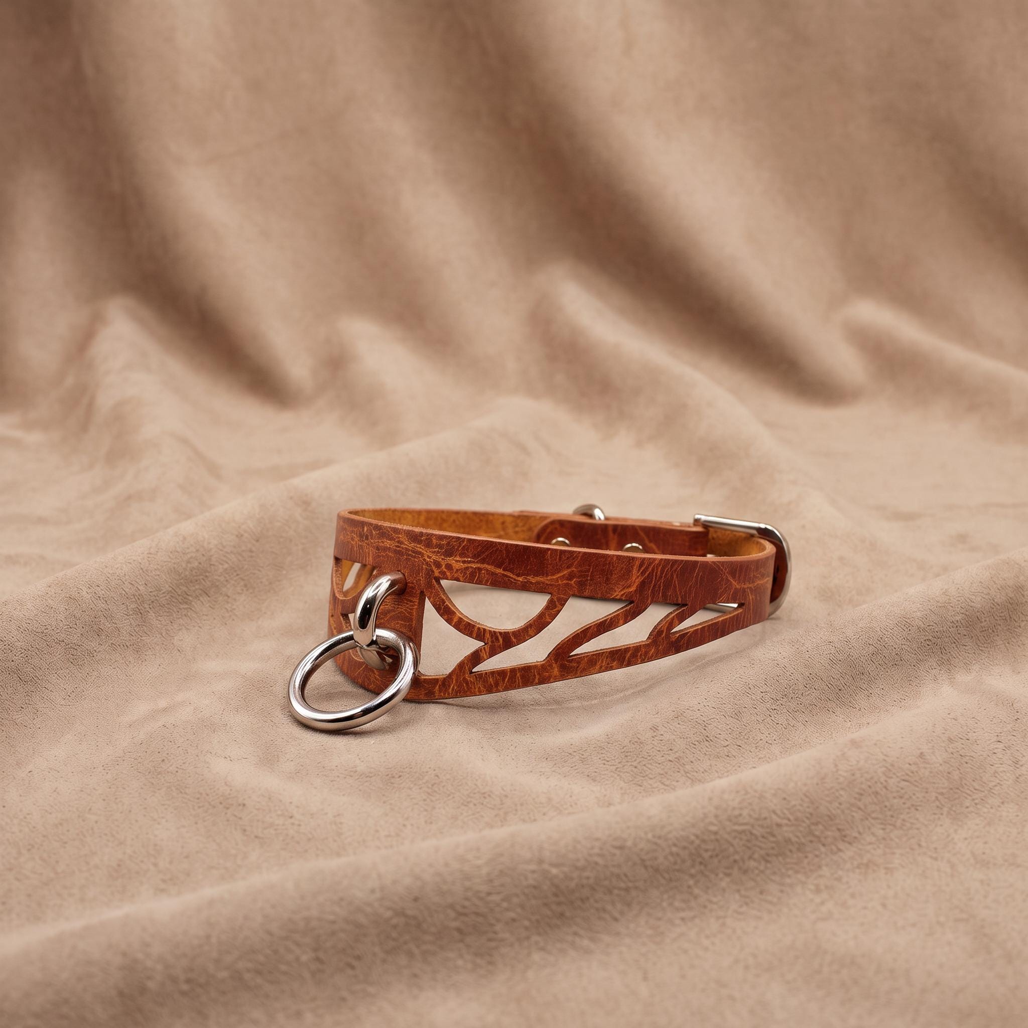 A brown leather choker necklace with a metal ring and buckle is displayed on a beige fabric background.