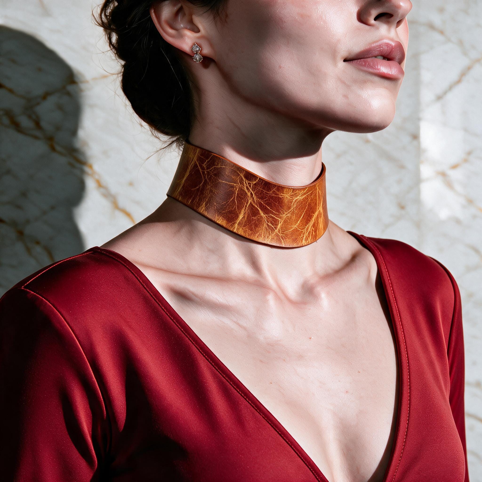 A woman wearing a red dress and a large, brown leather choker necklace stands against a marble wall.