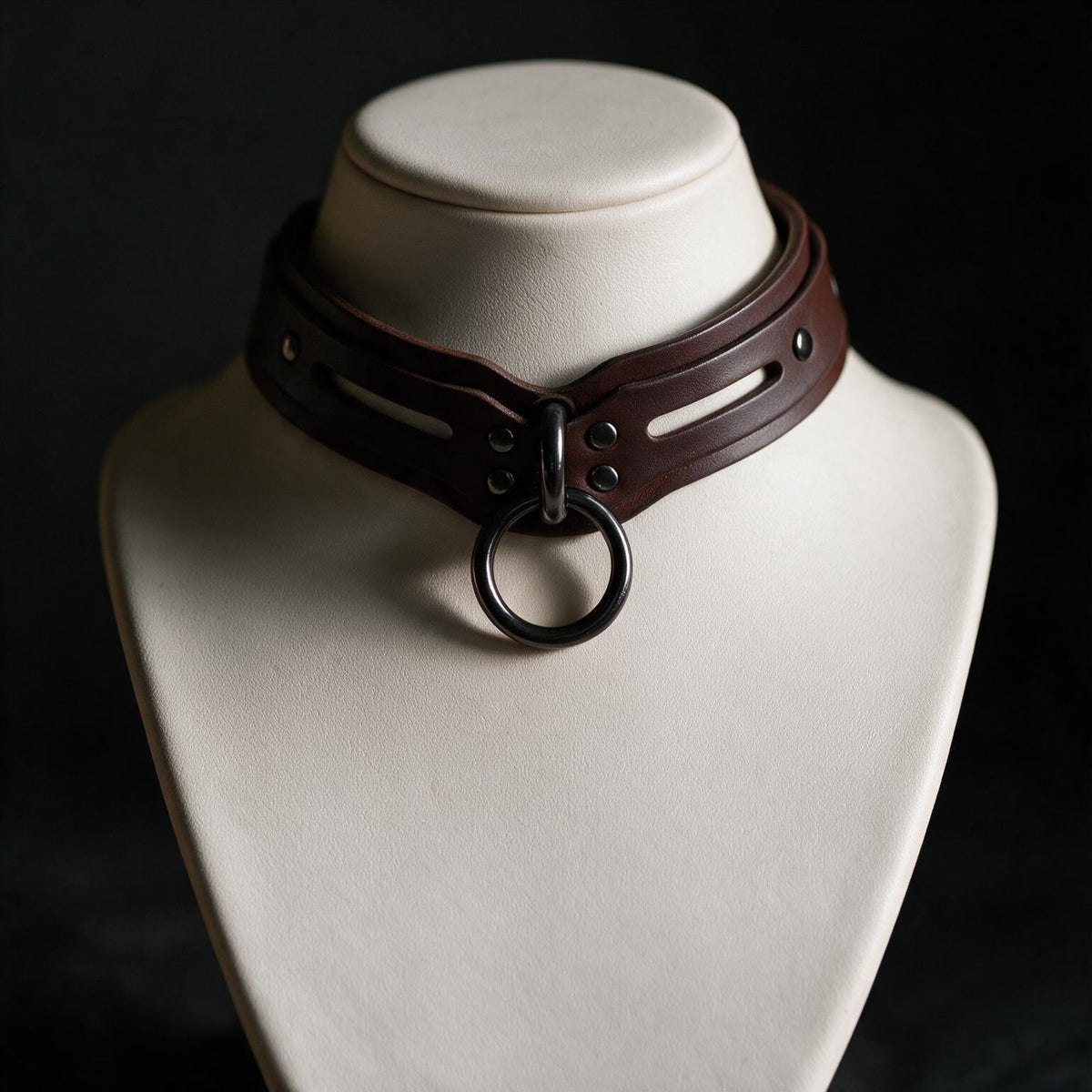 A close-up view of a leather choker necklace with a metal ring on the front, displayed on a white bust form against a dark background.