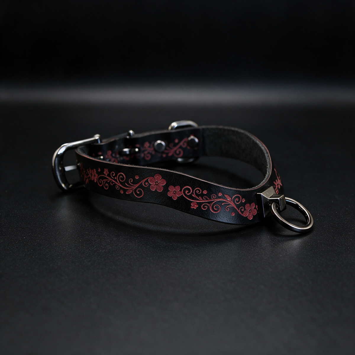 A black leather collar with a floral pattern and metal ring attached, resting on a dark surface.