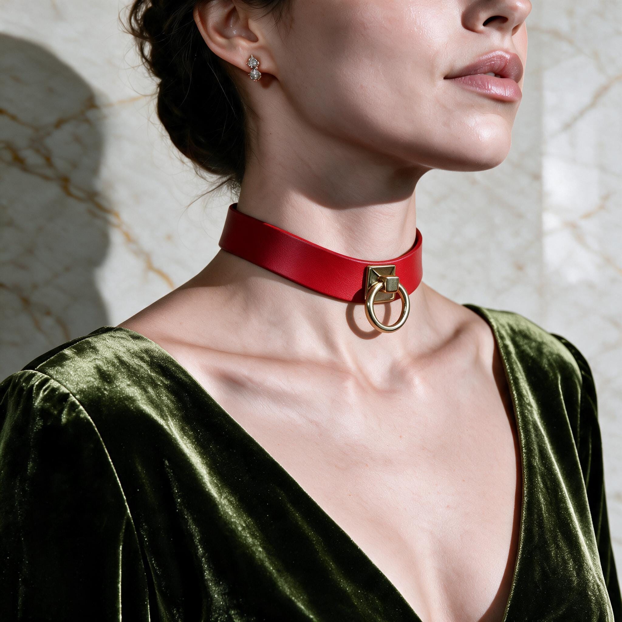 A woman wearing a red choker necklace with a gold ring on it, and a green velvet dress, posing against a marble wall.
