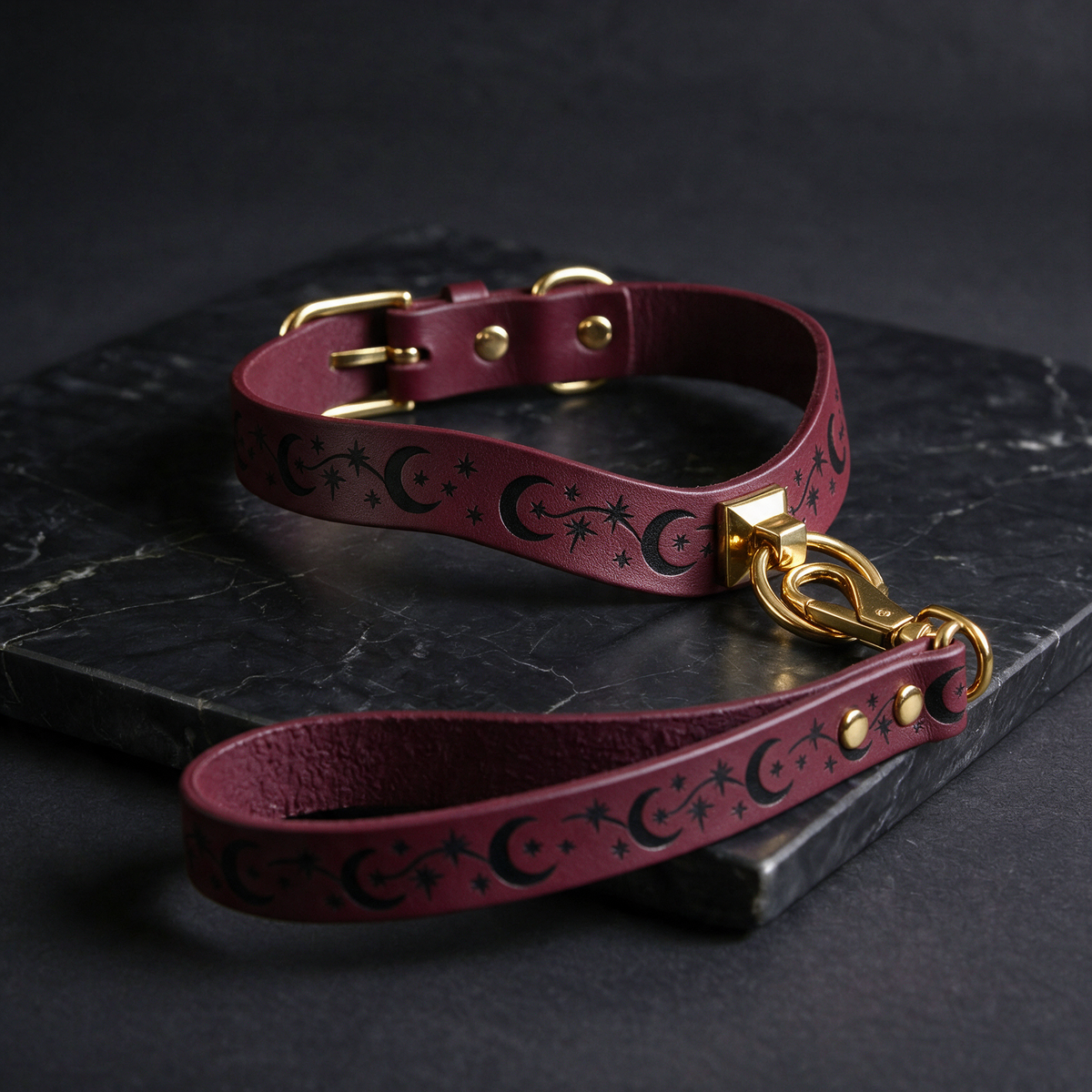 A burgundy leather dog collar with a starry night sky design and a gold-colored buckle is displayed on a black marble surface.