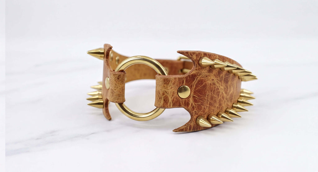 A brown leather bracelet with gold spikes protruding from it, resting on a white marble surface.