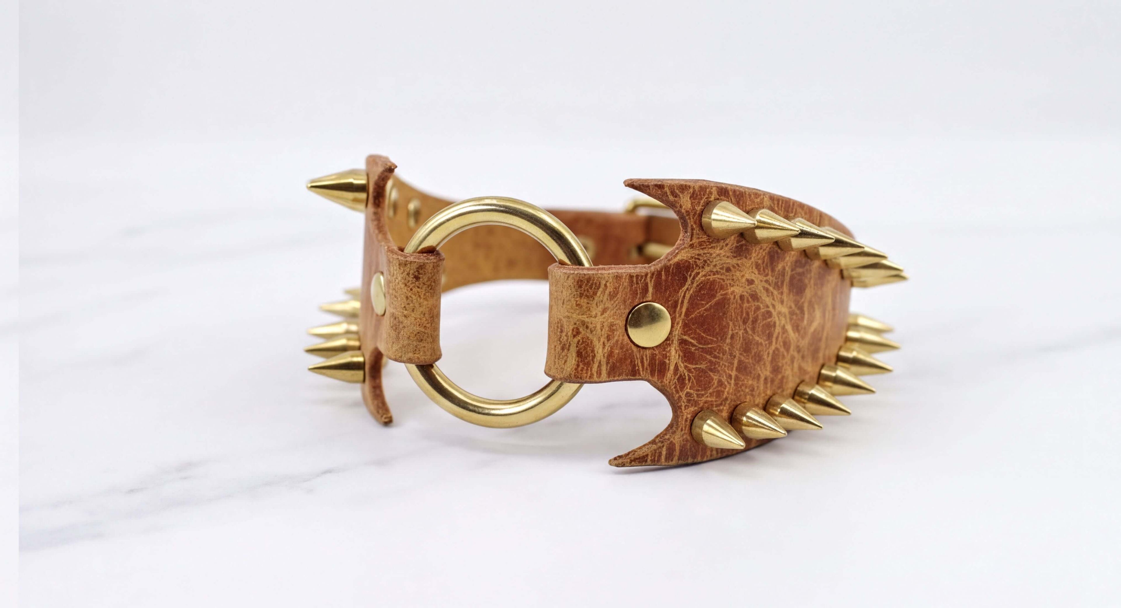 A brown leather bracelet with gold spikes protruding from it, resting on a white marble surface.