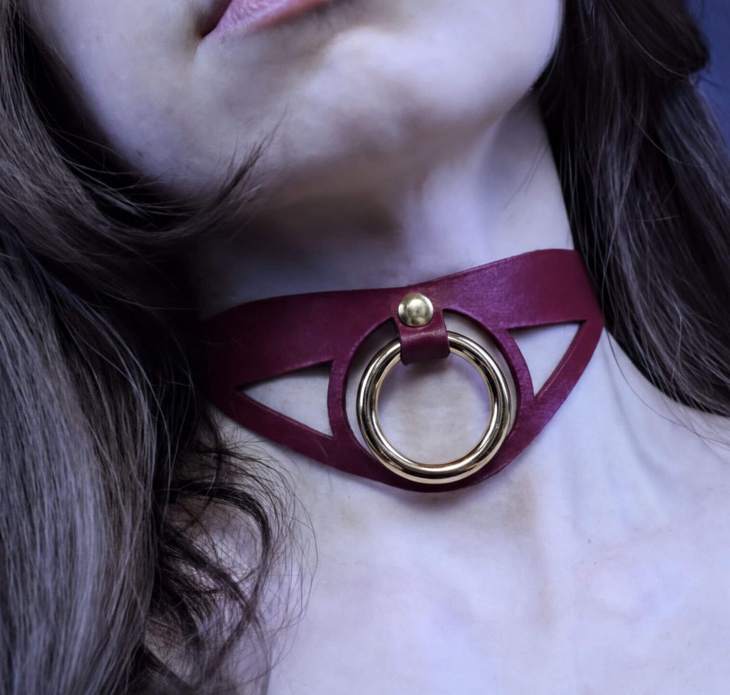 A close-up of a woman's neck with a burgundy choker necklace featuring a gold ring.