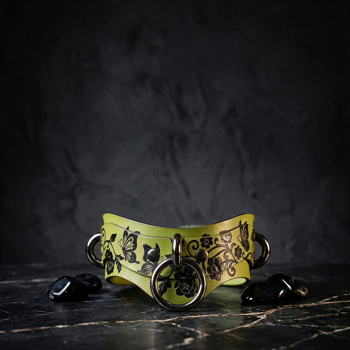 A yellow leather collar with black floral designs and metal rings is displayed on a dark surface, accompanied by small black stones.