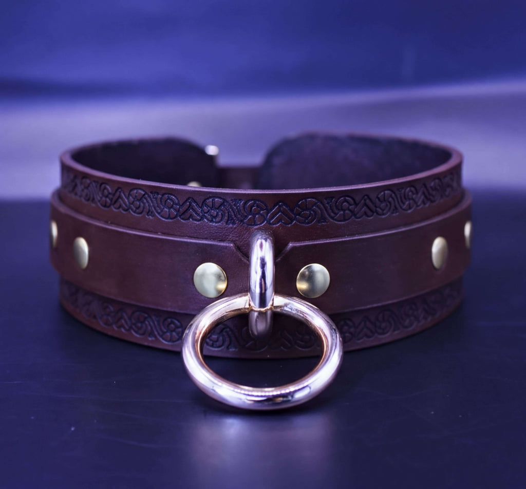 A brown leather choker collar with a metal ring and decorative elements, displayed on a dark surface against a blue background.