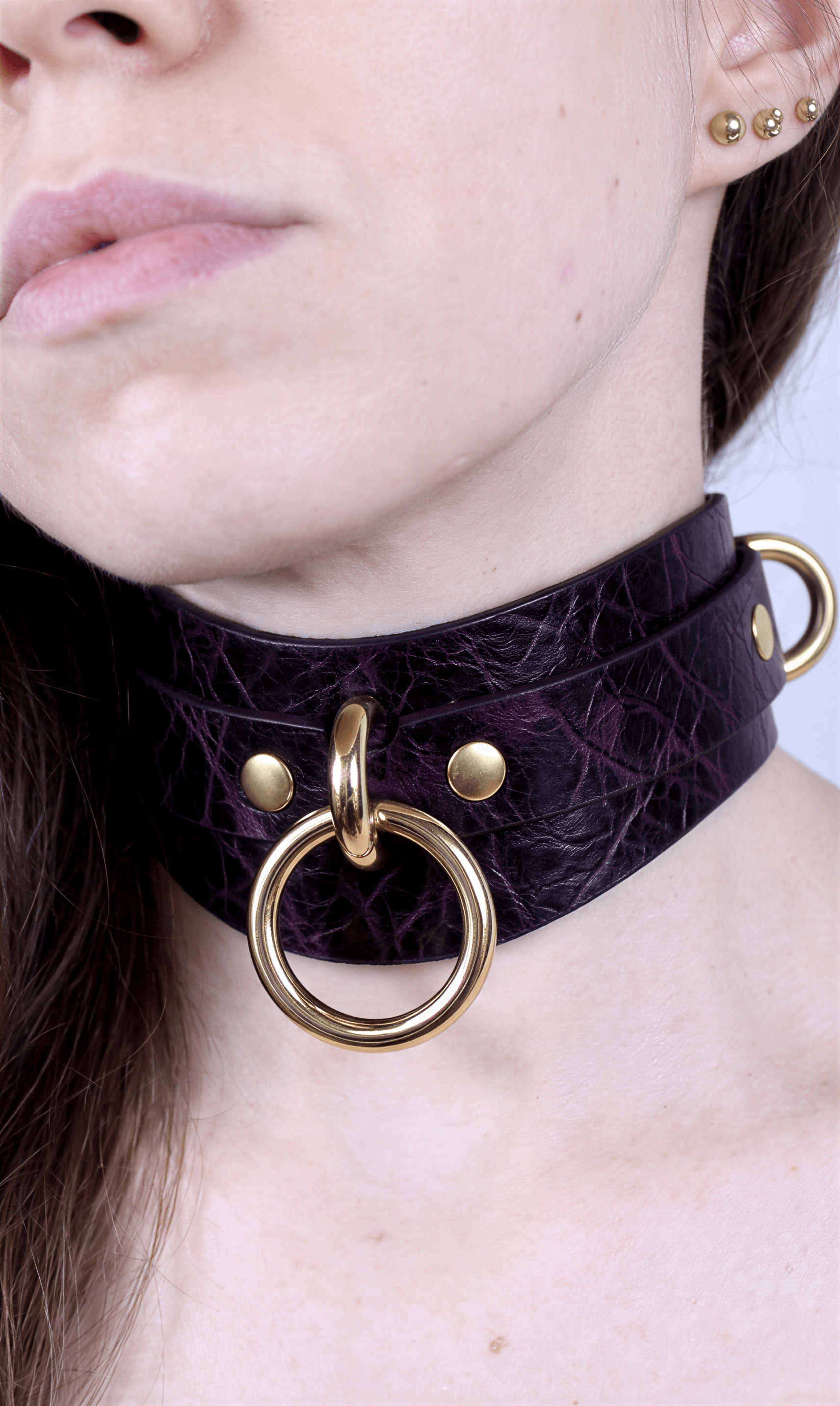 A close-up of a person's neck and chin area, featuring a dark purple choker necklace with a gold ring and two gold-colored rings on either side of the necklace.
