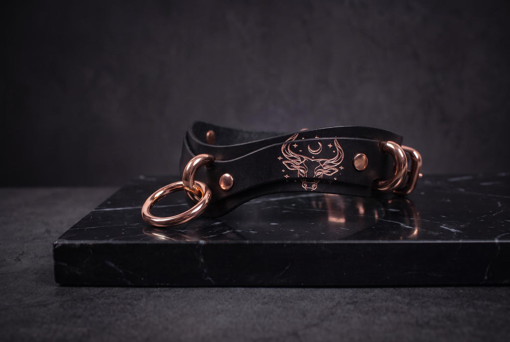A black leather collar with a metallic buckle and a decorative emblem is displayed on a black marble surface.