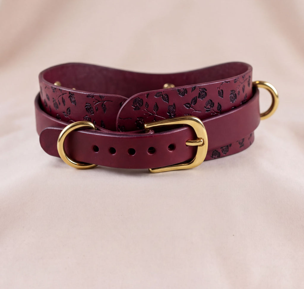 A burgundy leather belt with a floral pattern and gold-colored buckles, displayed on a light-colored fabric background.