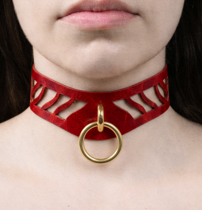 A close-up of a person's neck and chest area wearing a red choker necklace with a gold ring.