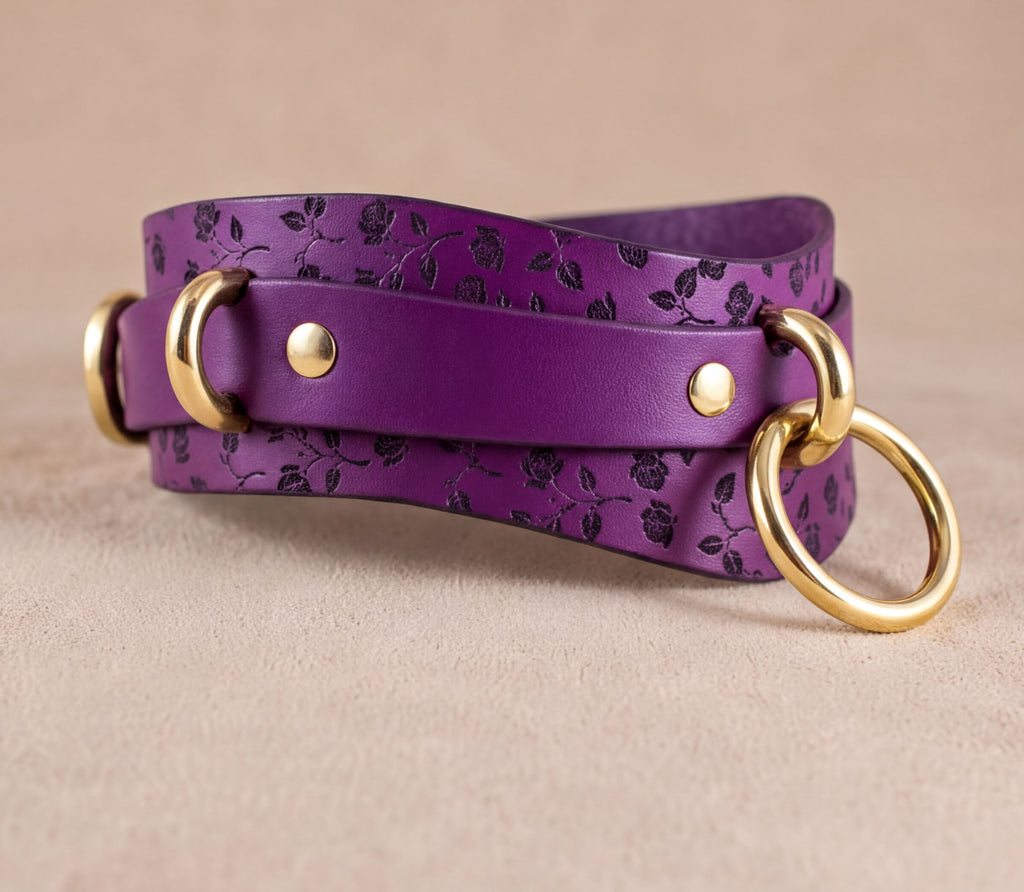 A purple leather strap with a floral pattern and two gold rings on the ends, resting on a beige surface.