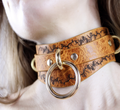A close-up of a leather choker necklace with a gold ring and decorative elements.